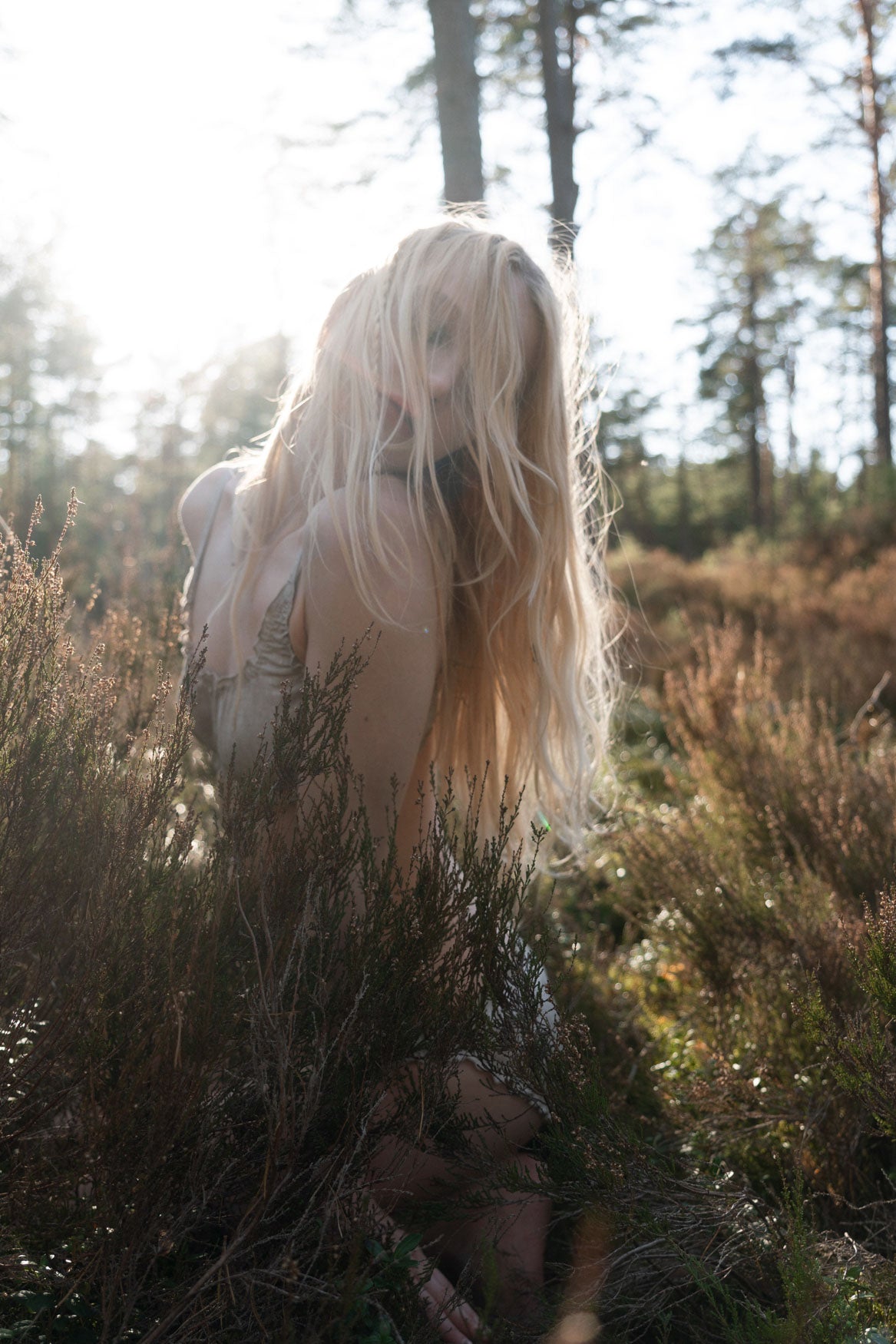 Person with long blonde hair standing in a forest with sunlight filtering through the trees.